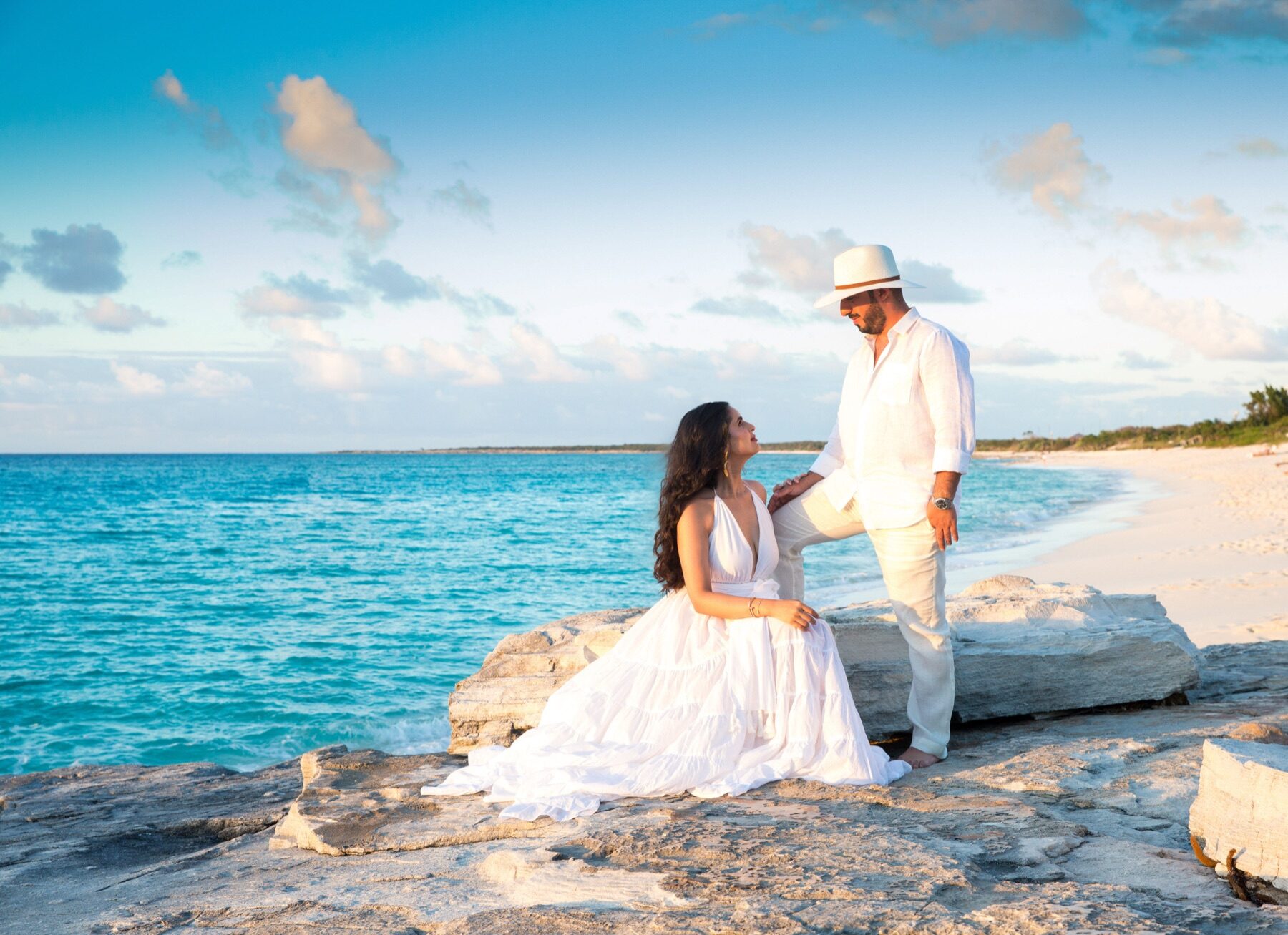 Couple in white facing each other on a rocky point above turquoise water
