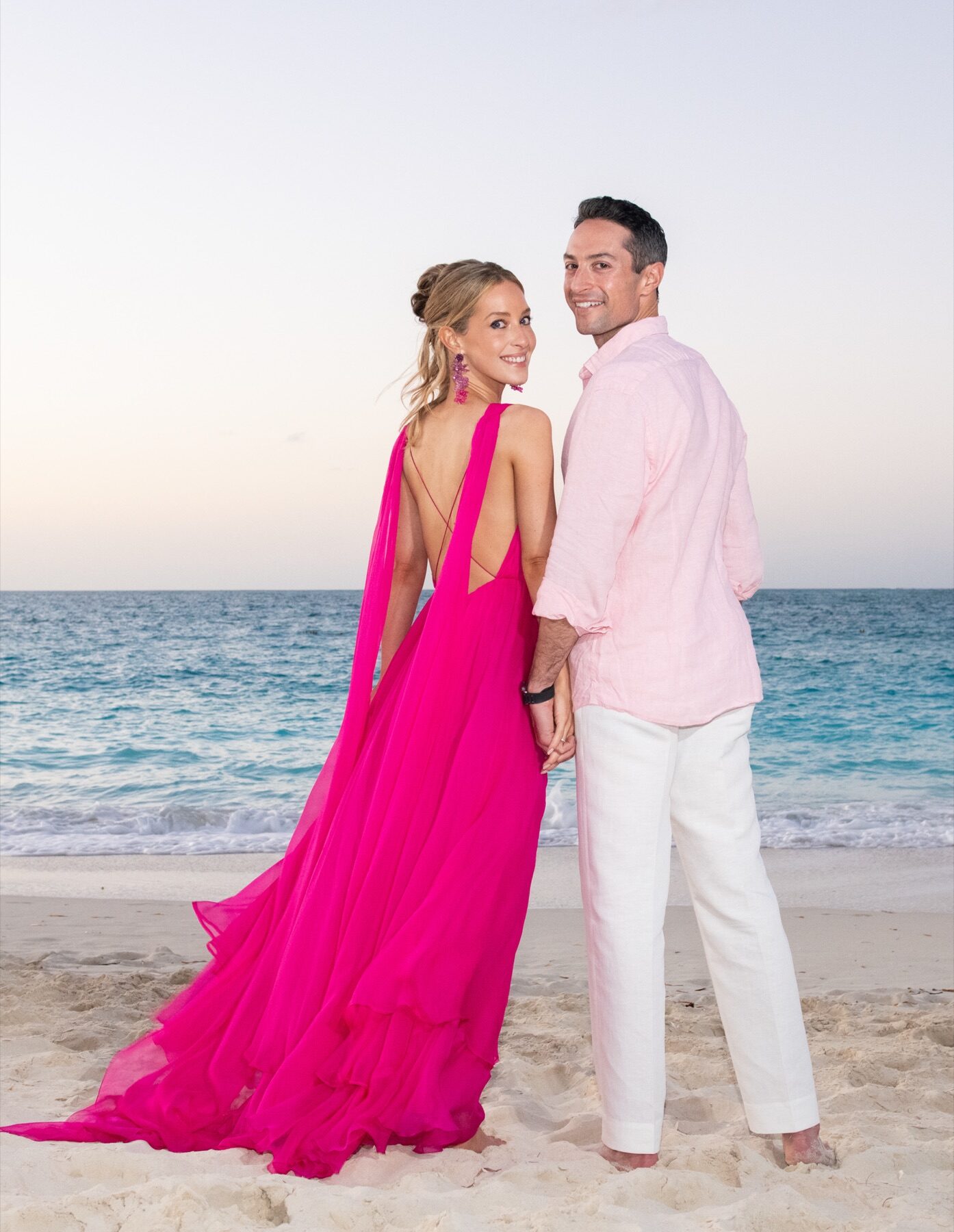 Couple in pink and white standing on the beach, woman glancing back over her shoulder