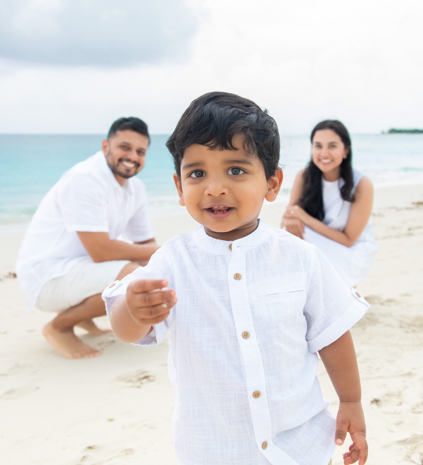 Toddler boy pointing at the camera as his parents crouch behind him, Turks and Caicos