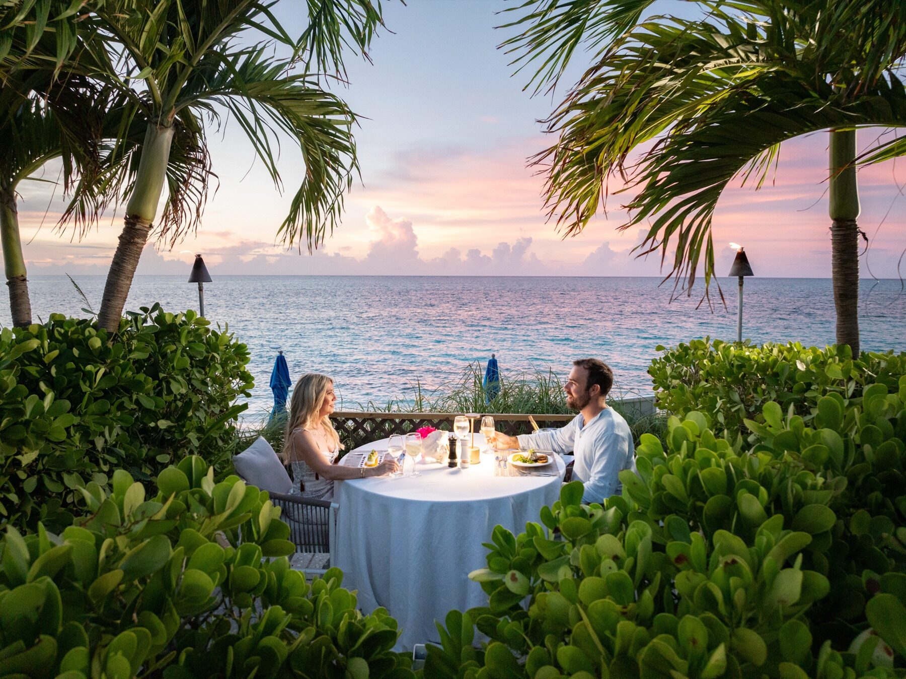 Couple sharing a candlelit dinner table beneath palm trees at sunset