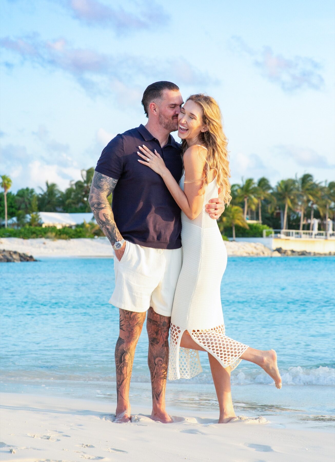 Tattooed man kissing his fiancee's cheek while she kicks up a bare foot in the sand