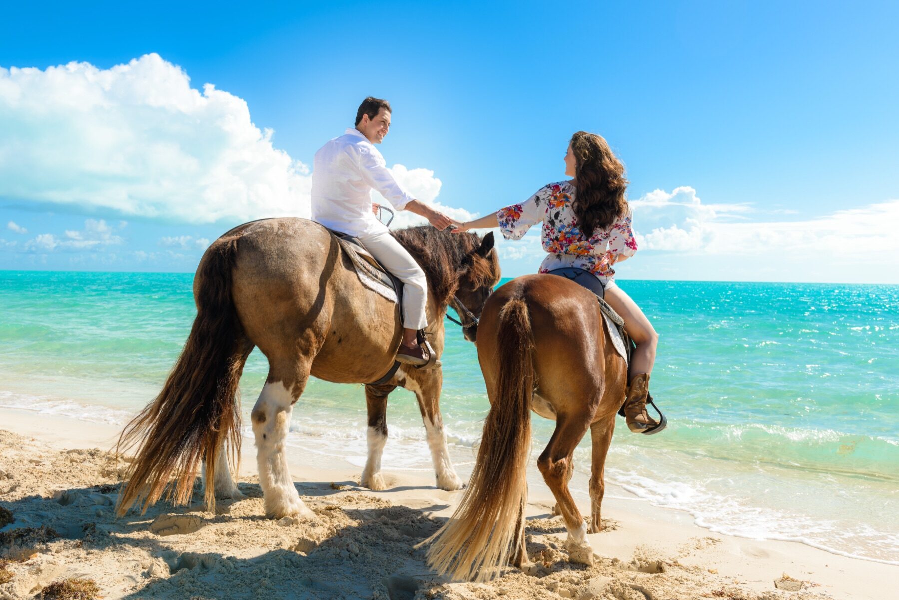 Couple holding hands while riding horses along the shoreline