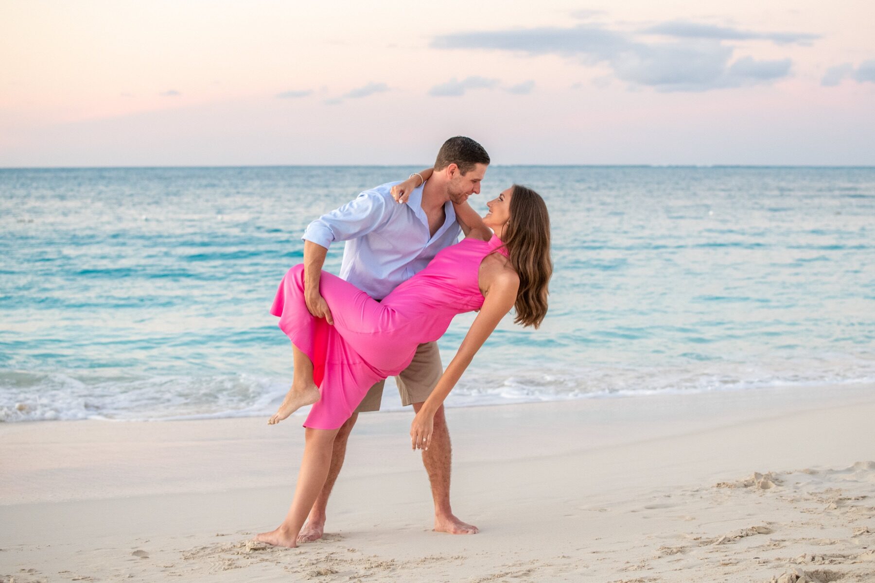 Man dipping his newly engaged partner in a pink dress on the beach