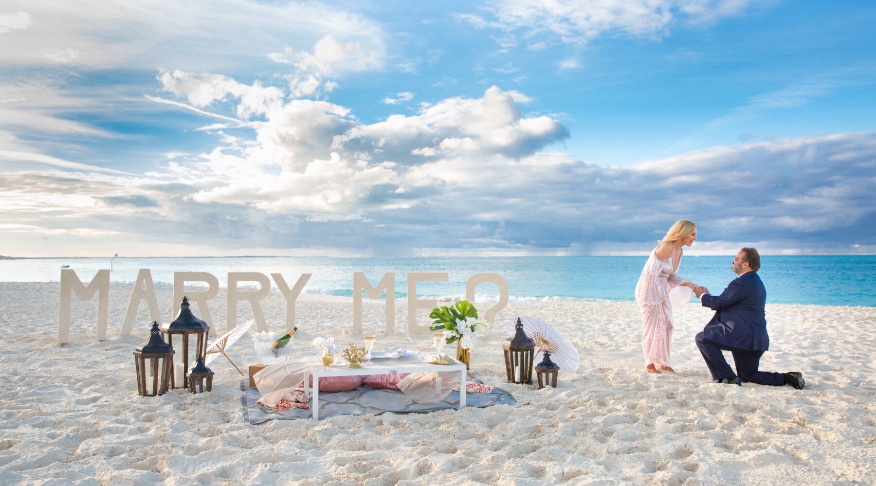 Man on one knee proposing beside giant marry me letters set up on the sand
