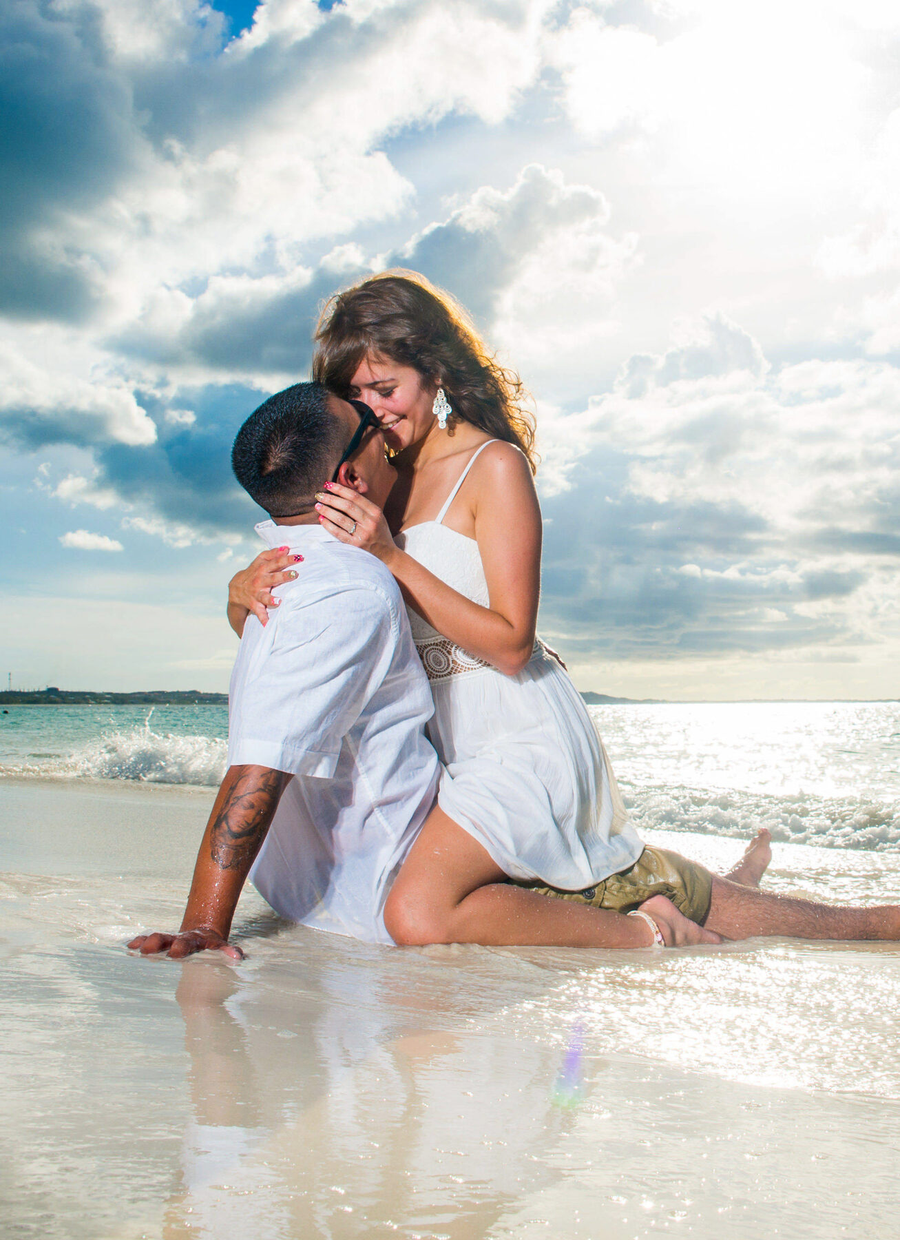 Newly engaged couple embracing at the water's edge under a bright, cloud-streaked sky