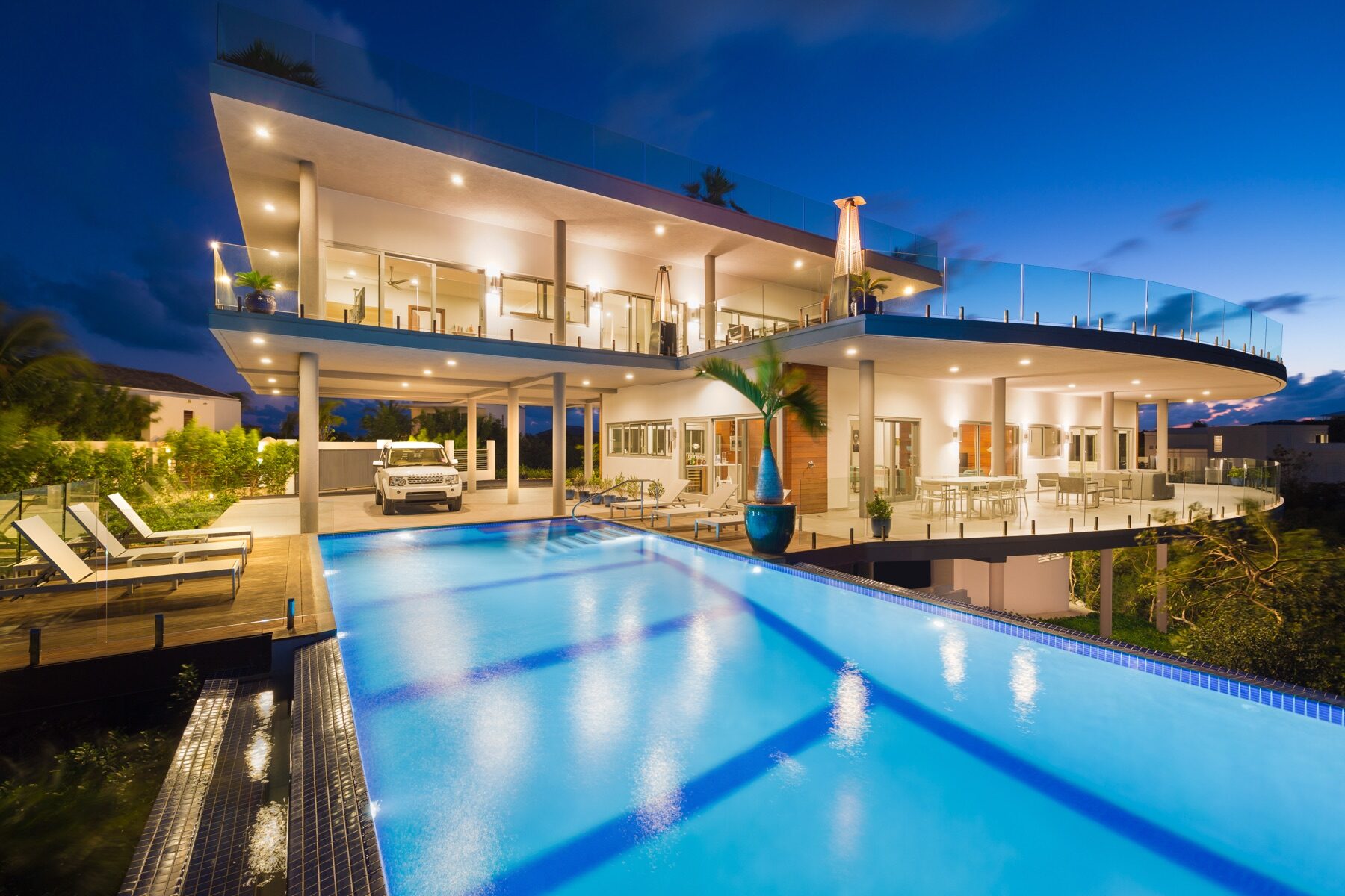 Modern villa at dusk with a long lit pool beside a covered carport