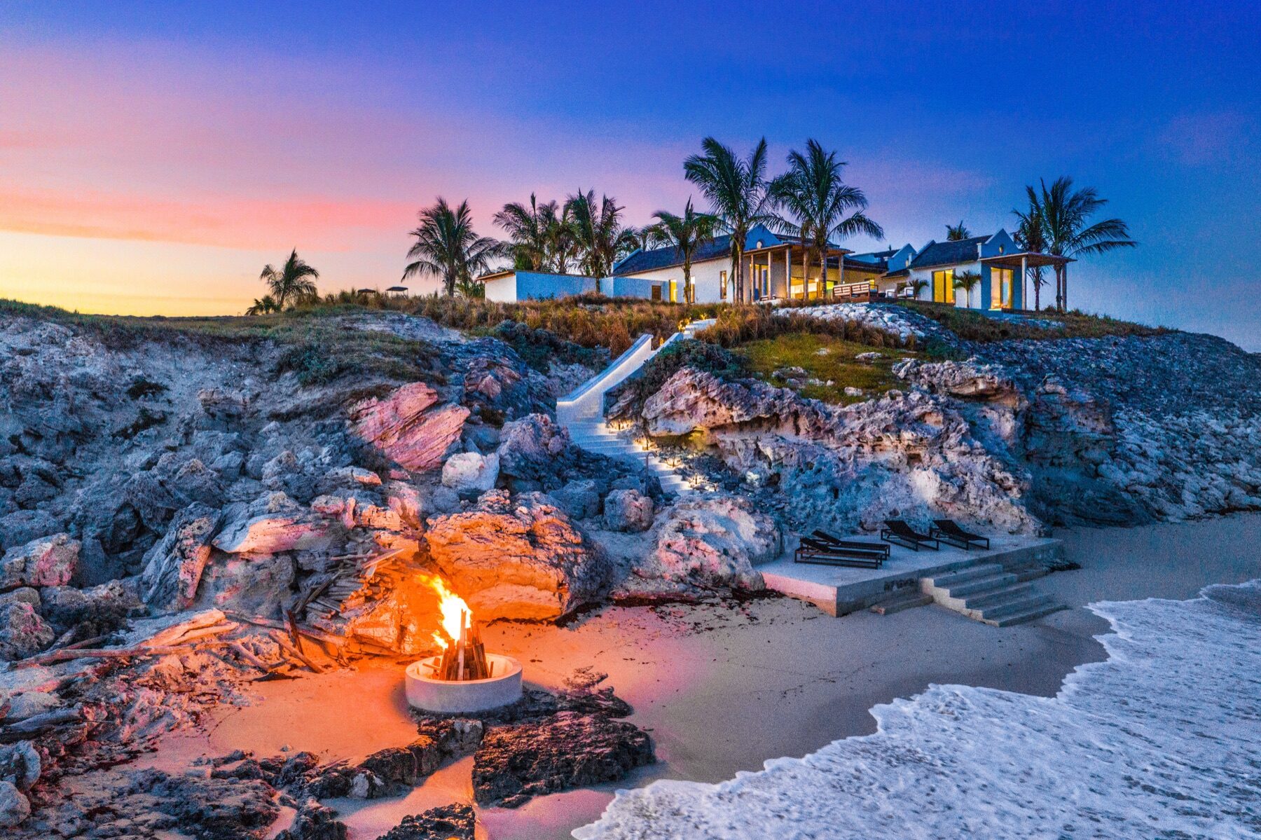Bonfire glowing on a rocky beach beneath a hillside villa at twilight