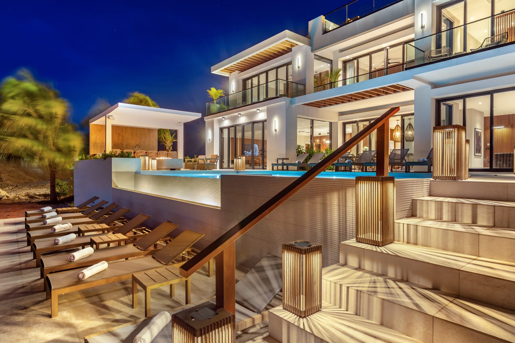 Villa exterior at night with an infinity pool and wooden staircase lighting