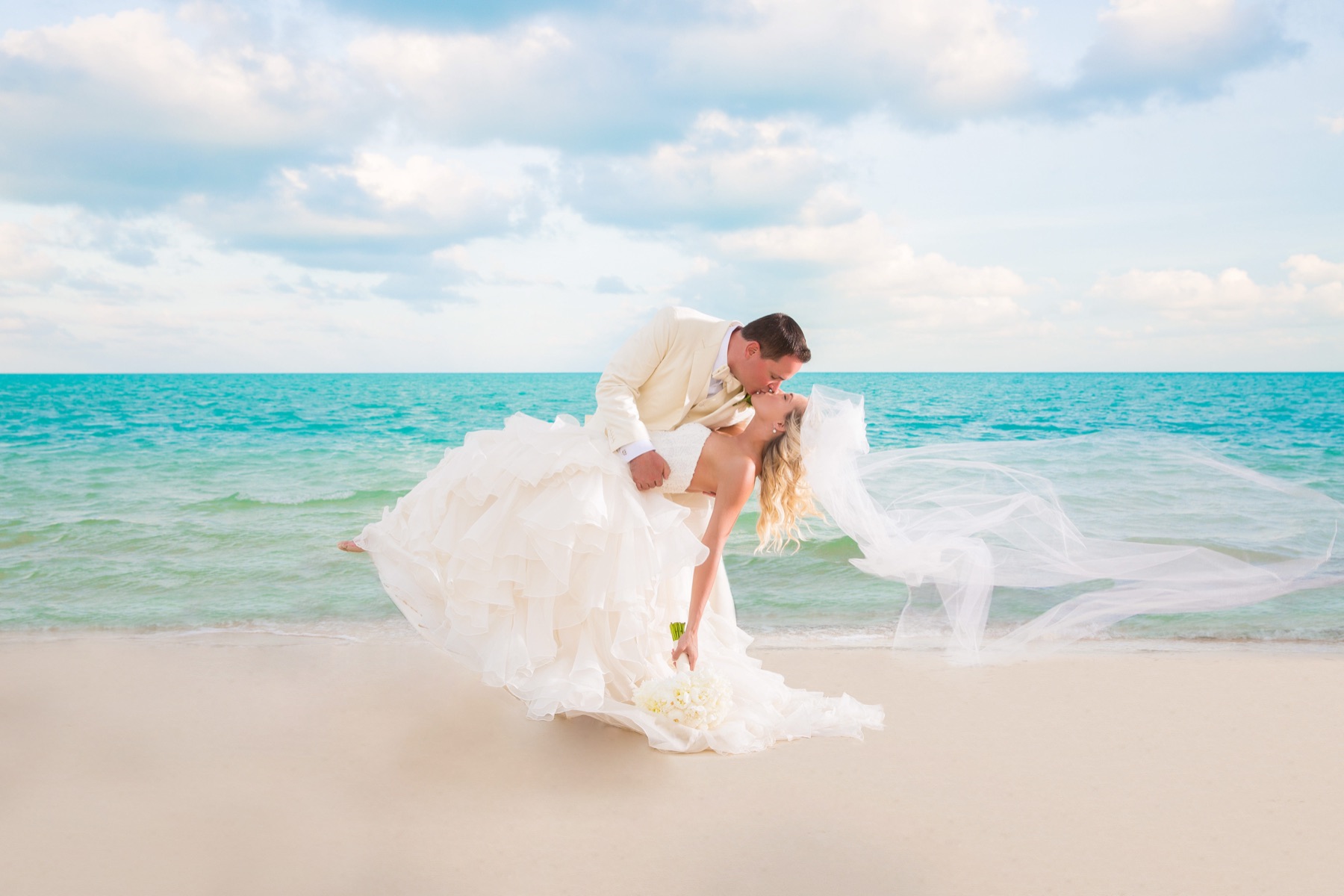Groom dipping bride for a kiss on the beach as her veil catches the wind