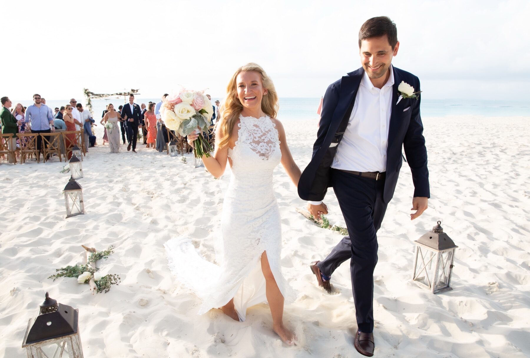 Bride and groom walking hand in hand past lanterns lining the sand aisle