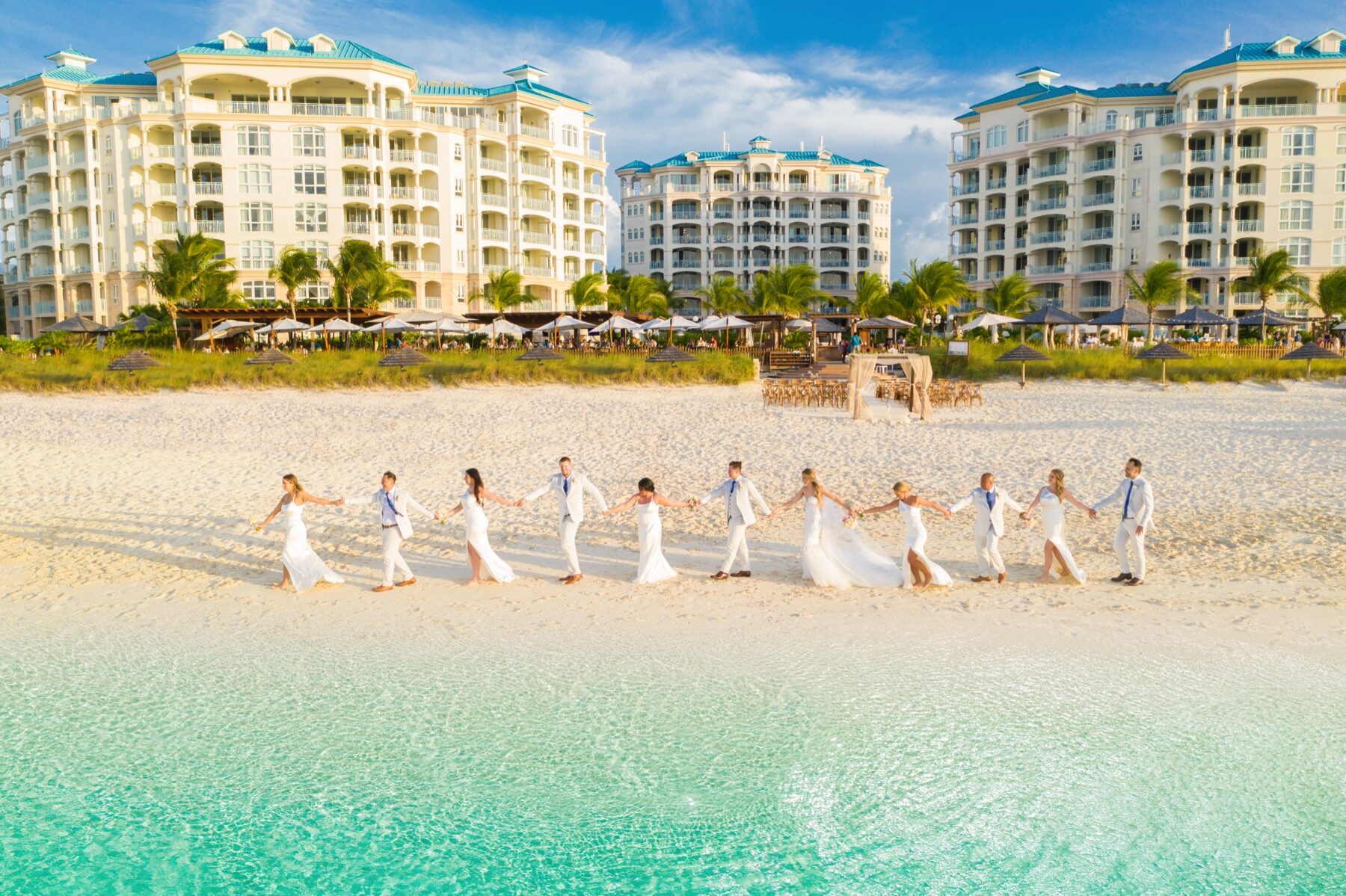 Wedding party in white walking hand in hand along the shallow turquoise shoreline
