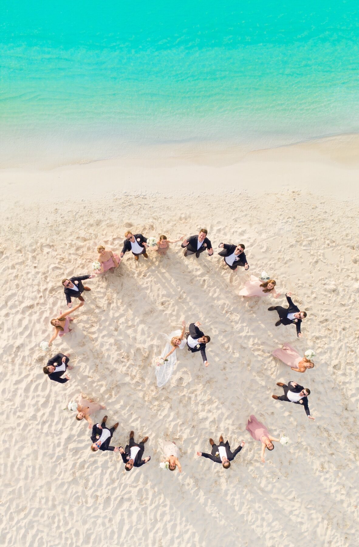 Aerial view of a wedding party forming a circle around the couple on the sand