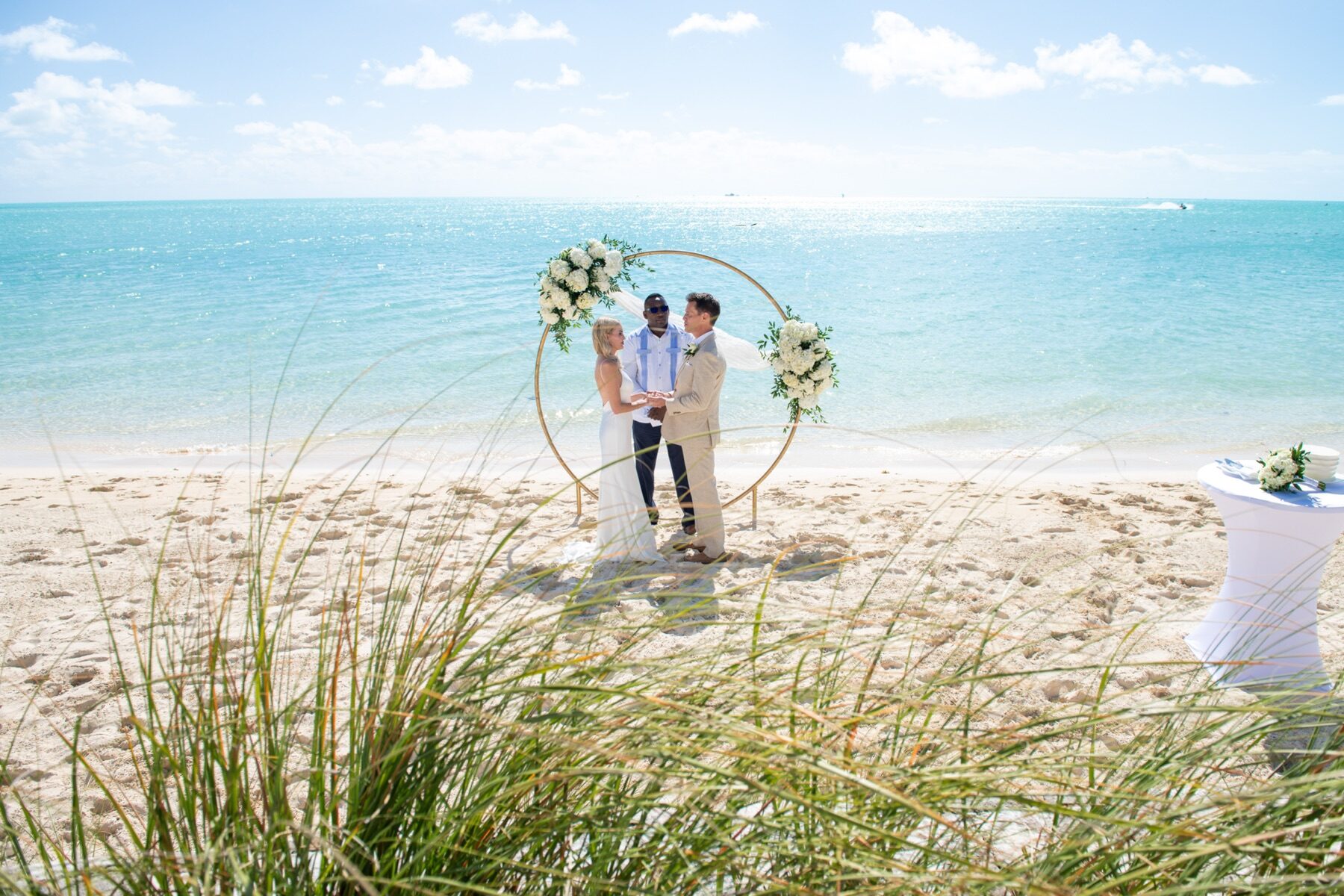 Officiant marrying a couple under a round floral arch with beach grass in the foreground