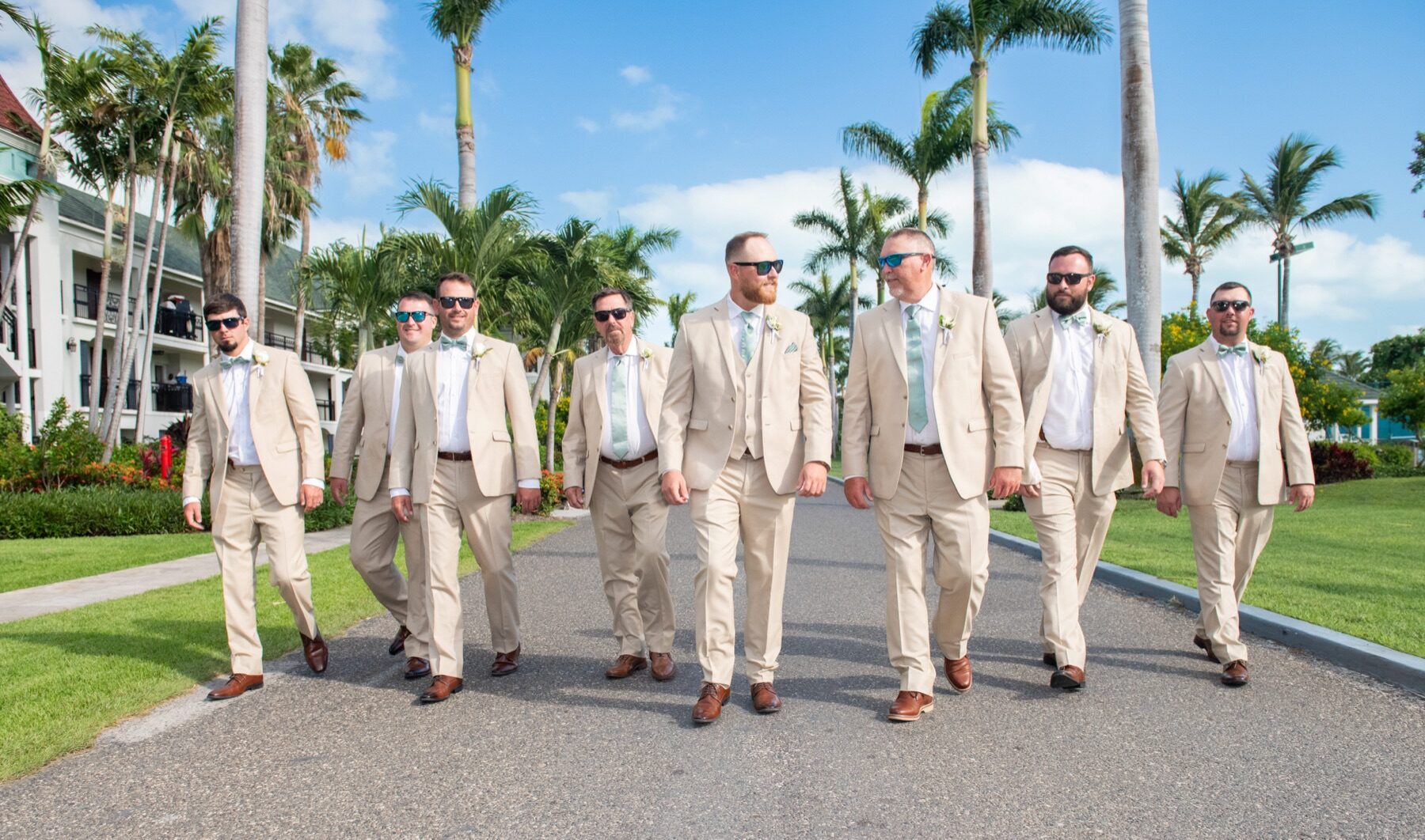 Groomsmen in tan suits and sunglasses walking down a Turks and Caicos palm lined path